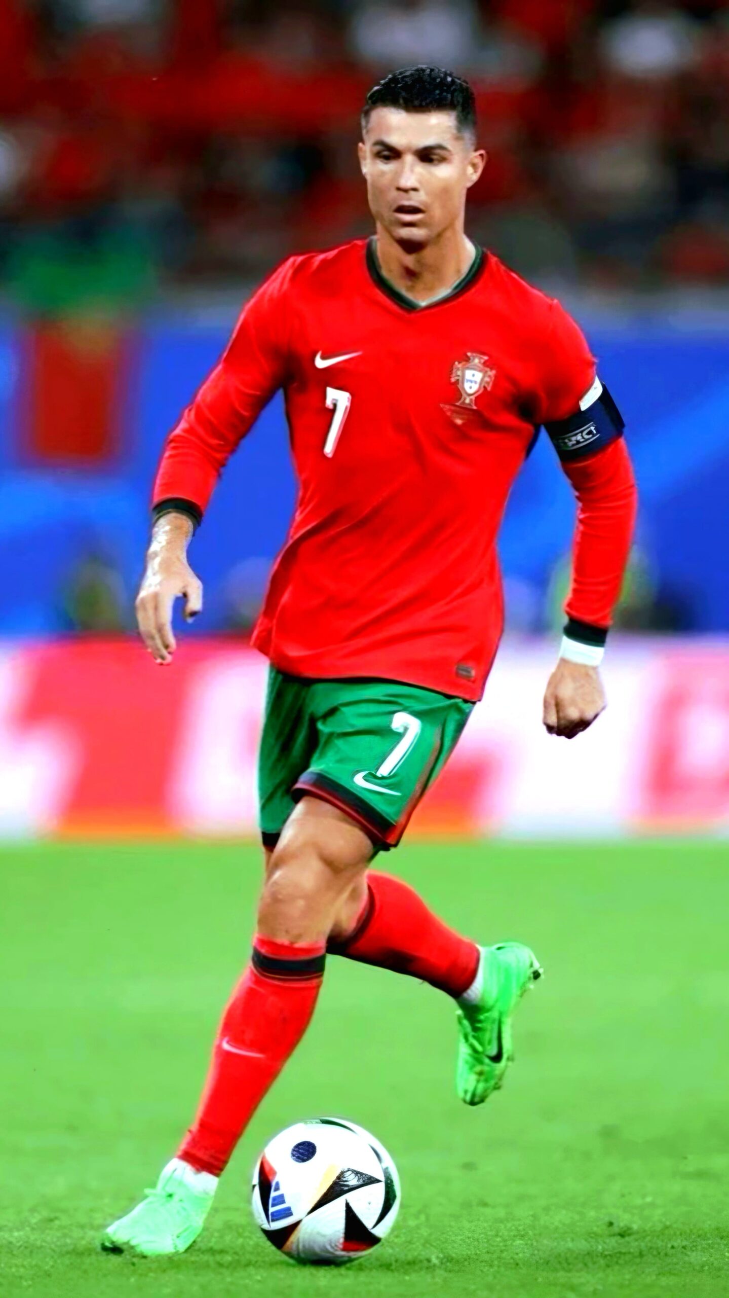 A male soccer player in a red and green Portugal uniform with the number 7 dribbles a soccer ball on the field. He wears a captain's armband and green cleats, with a stadium crowd blurred in the background, streaming live on Malta IPTV.