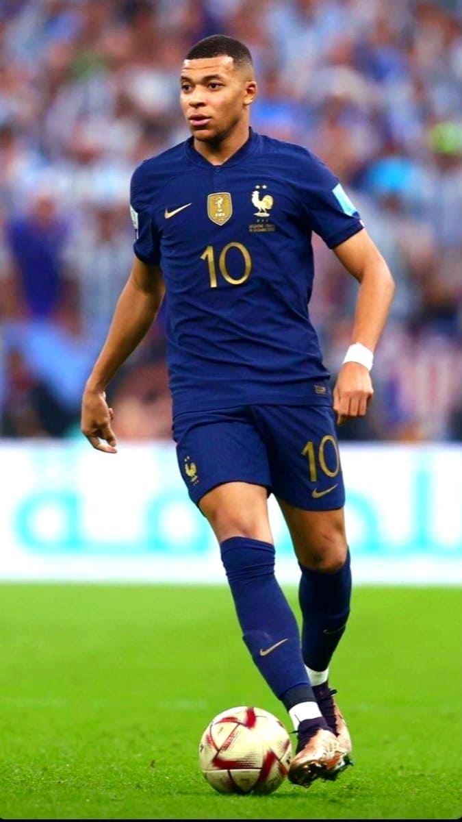 A male soccer player in a dark blue France national team uniform with the number 10 is dribbling a ball on a grass field during a match, captured vividly for fans watching via Malta IPTV. The background is blurred with spectators.