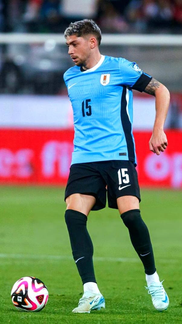 A soccer player in a light blue Uruguay jersey with the number 15 controls a colorful ball on the field during a match. Fans watching via Malta IPTV see him focused, with one leg bent and the other extended.