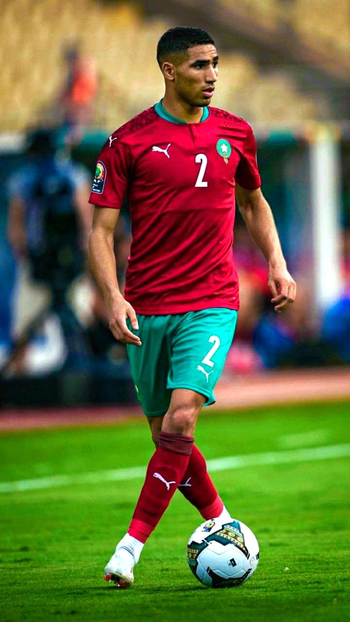 A male soccer player in a red and green Morocco national team uniform with the number 2 dribbles a white and blue ball on the field during a match, broadcast live via Malta IPTV.