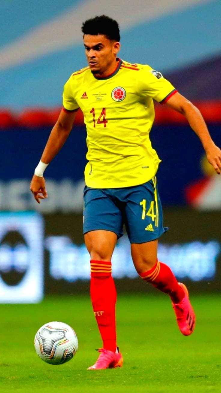 A soccer player in a yellow Colombia jersey with the number 14 is running on the field, controlling the ball. Shown live on Malta IPTV, he sports blue shorts, red socks, and pink cleats, his expression focused during the match.